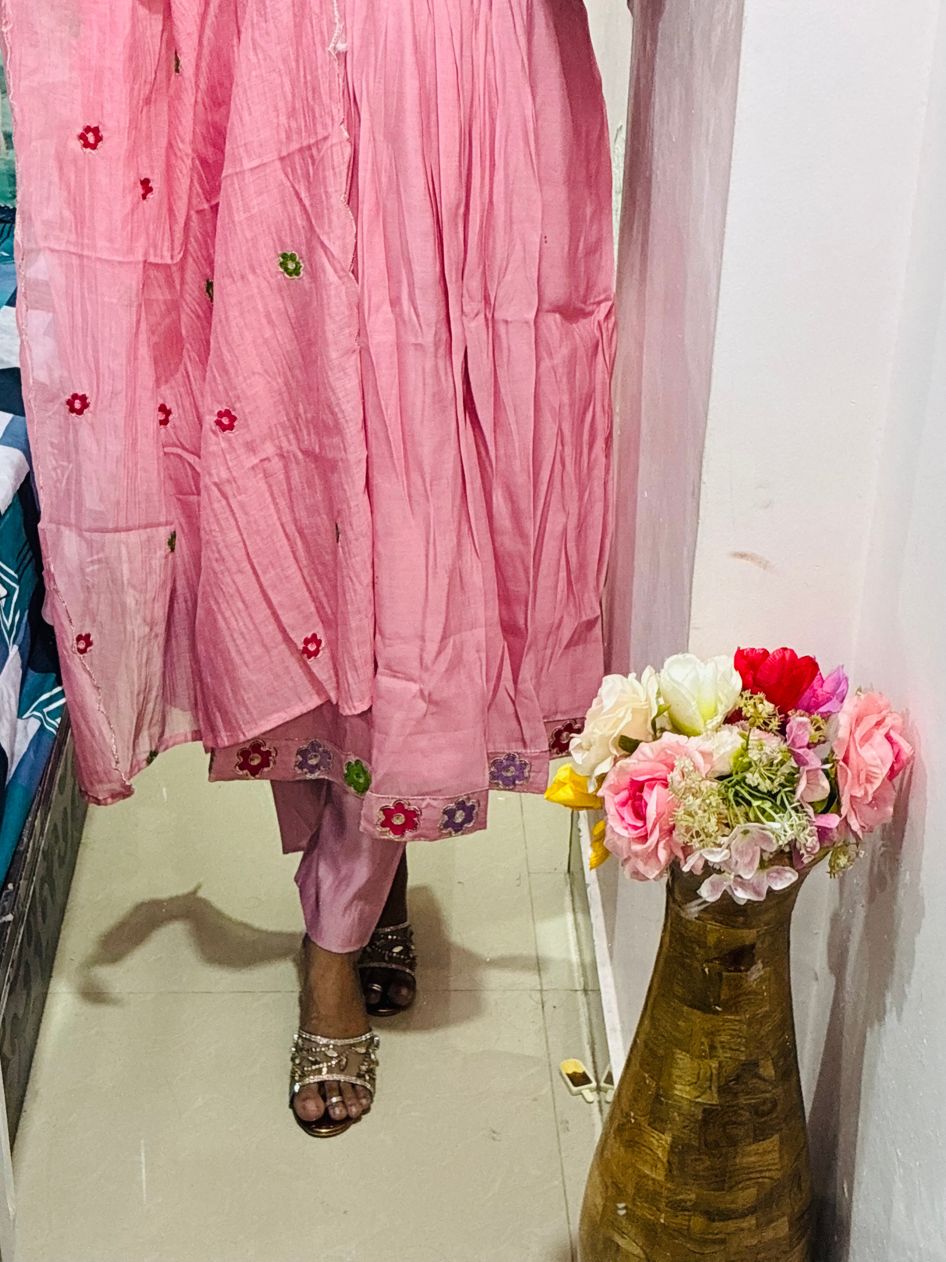 Pink traditional outfit with floral details on a person, standing next to a vase with flowers.